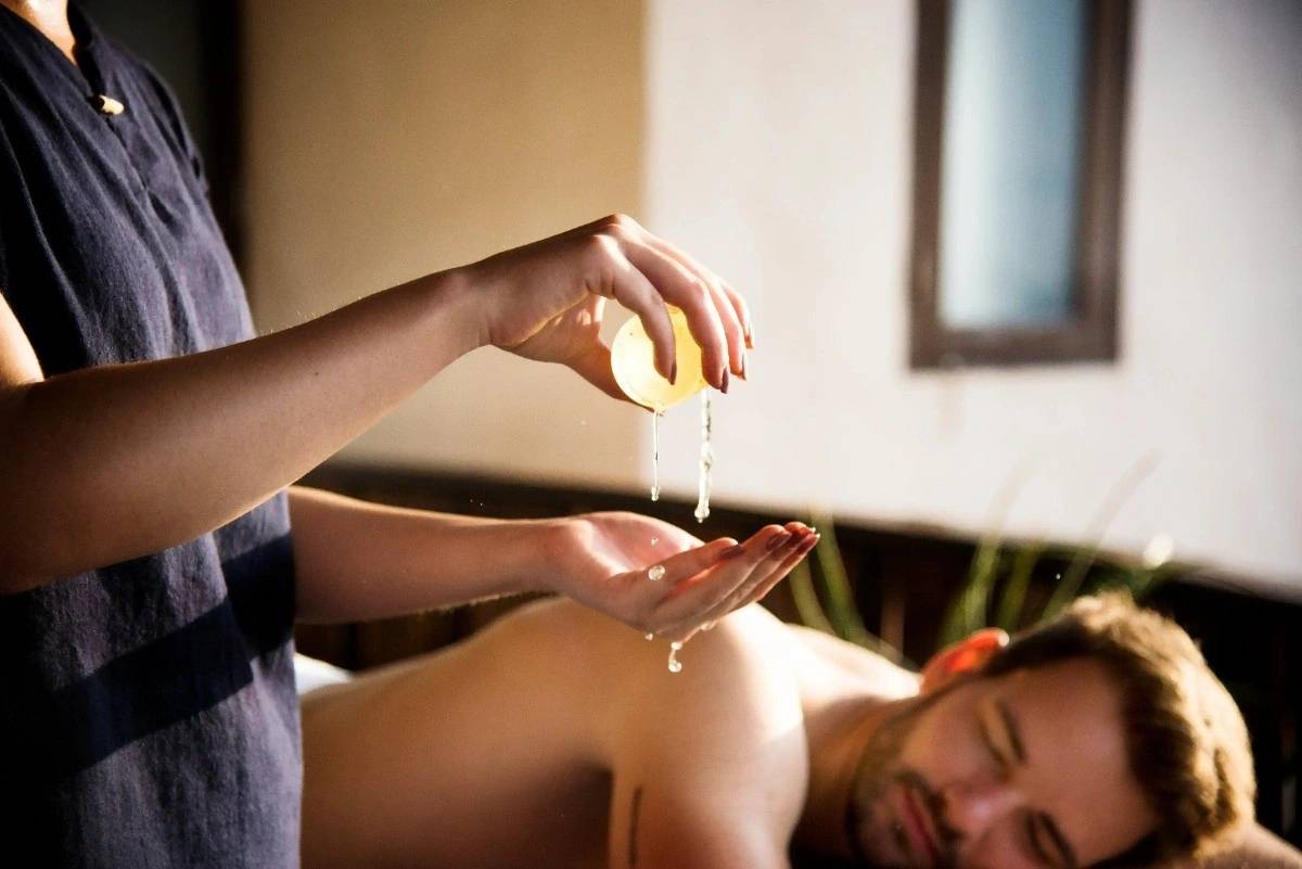 A therapist pouring warm aromatic oil into her palm above a guest's back during a massage