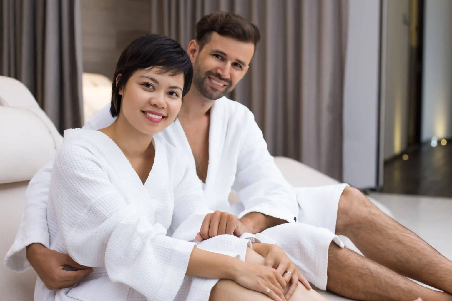 A smiling couple in white bathrobes relaxing together in a softly lit spa suite
